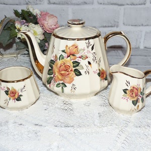 May include: A vintage tea set with a cream-coloured base and gold trim. The teapot, sugar bowl and creamer feature a floral pattern with yellow roses and green leaves.