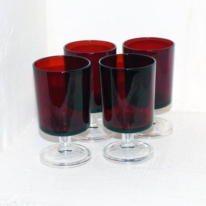 May include: Four red glass goblets with clear stems and bases. The goblets are arranged in a row on a white surface.