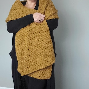 May include: Mustard yellow crocheted shawl with a textured, open weave design. The shawl is draped over a person wearing a black outfit. The shawl appears soft and warm, suitable for cooler weather. The color is a rich, earthy tone.