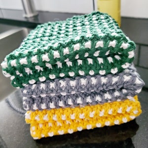 Pode incluir: Um conjunto de três panos de cozinha de malha em verde, cinza e amarelo com detalhes brancos. Cada pano tem um padrão texturizado.