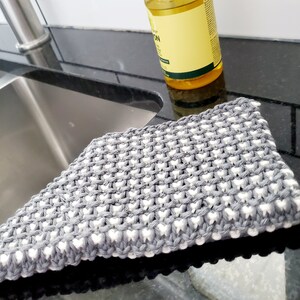 May include: A gray and white knitted dishcloth with a textured pattern. The dishcloth is lying on a black countertop.