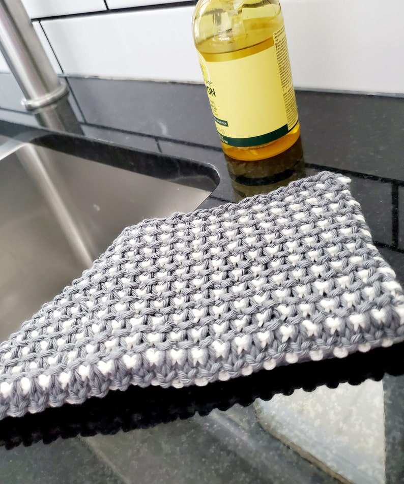 May include: A gray and white knitted dishcloth with a textured pattern. The dishcloth is lying on a black countertop.