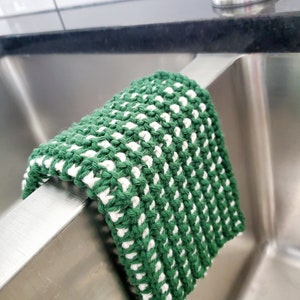 May include: A green and white knitted dishcloth hanging on the edge of a stainless steel kitchen sink.