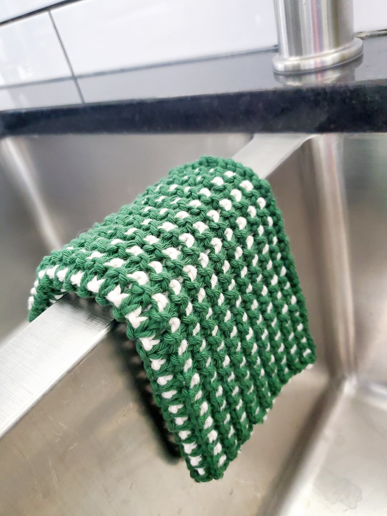 May include: A green and white knitted dishcloth hanging on the edge of a stainless steel kitchen sink.