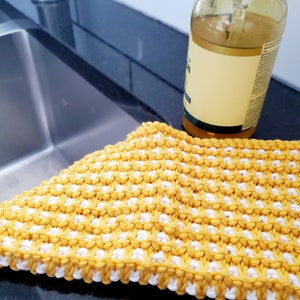 May include: A yellow and white knitted dishcloth with a textured pattern. The dishcloth is lying on a black countertop next to a clear glass bottle.