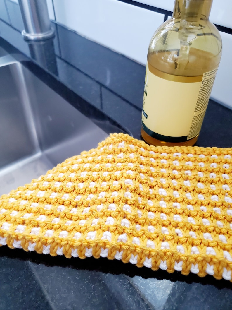 May include: A yellow and white knitted dishcloth with a textured pattern. The dishcloth is lying on a black countertop next to a clear glass bottle.