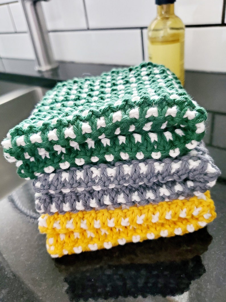 May include: A stack of three knitted dishcloths in green, gray, and yellow with white accents. Each dishcloth has a textured pattern.