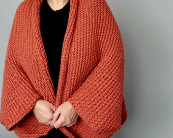 May include: An open-front, rust-colored knit cardigan with wide sleeves. The cardigan is made of a thick, textured yarn and has a relaxed fit. The model is wearing a black top underneath. The cardigan is a cozy and stylish outerwear option.