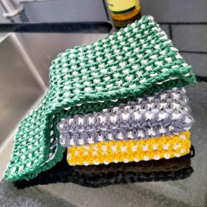 May include: A stack of three knitted dishcloths in green, gray, and yellow with white accents. The dishcloths are made with a textured stitch pattern.