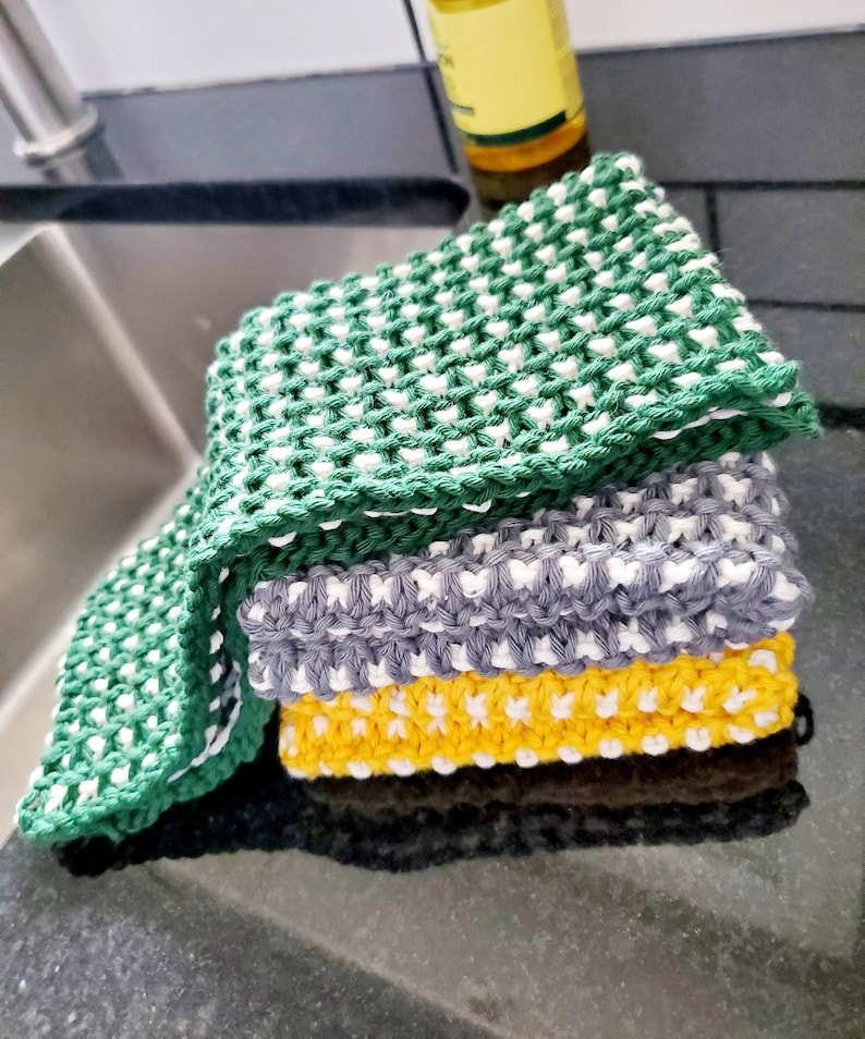 May include: A stack of three knitted dishcloths in green, gray, and yellow with white accents. The dishcloths are made with a textured stitch pattern.