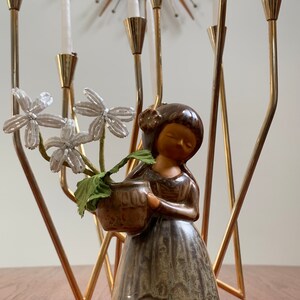 May include: A ceramic figurine of a woman holding a pot with white flowers. The figurine is standing in front of a gold metal candelabra with white candles.
