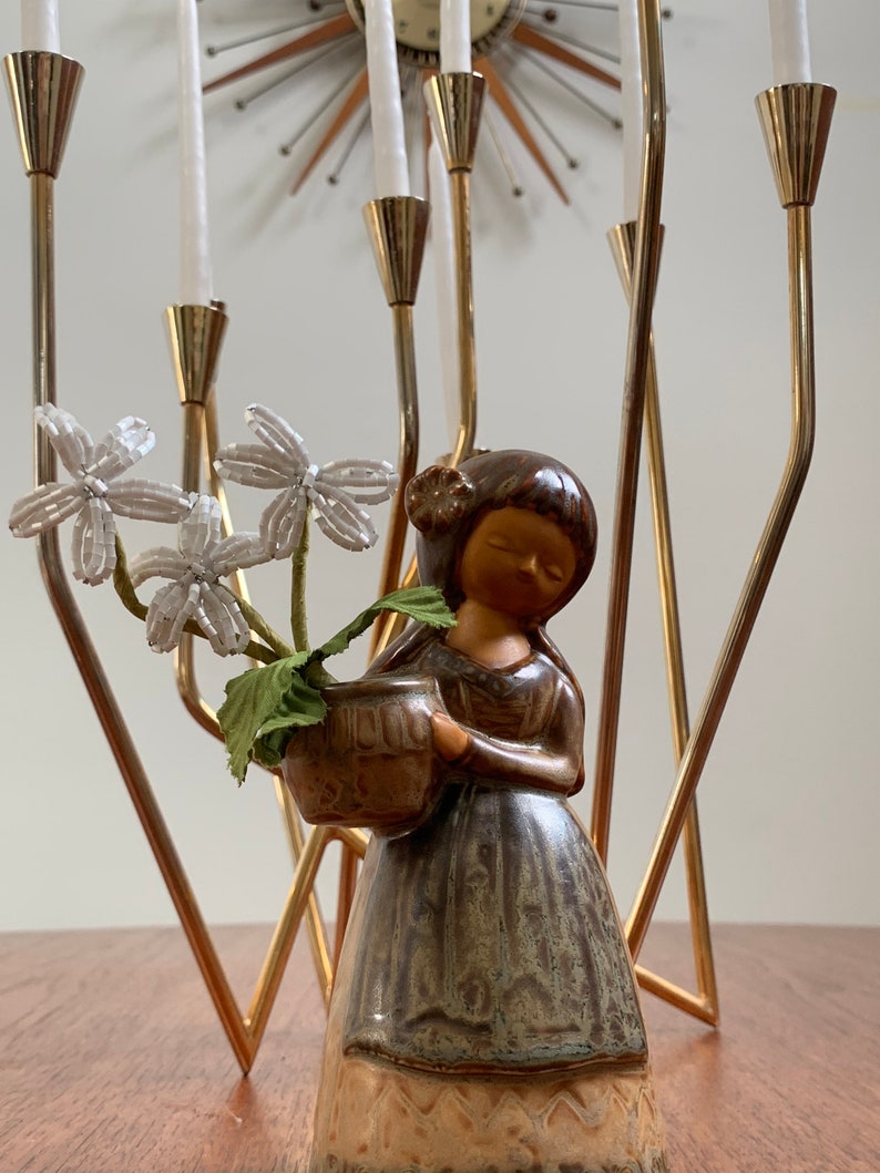 May include: A ceramic figurine of a woman holding a pot with white flowers. The figurine is standing in front of a gold metal candelabra with white candles.