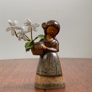 May include: A ceramic figurine of a girl holding a basket of white flowers. The girl is wearing a brown dress with a green and brown pattern. The figurine is made in a rustic style.