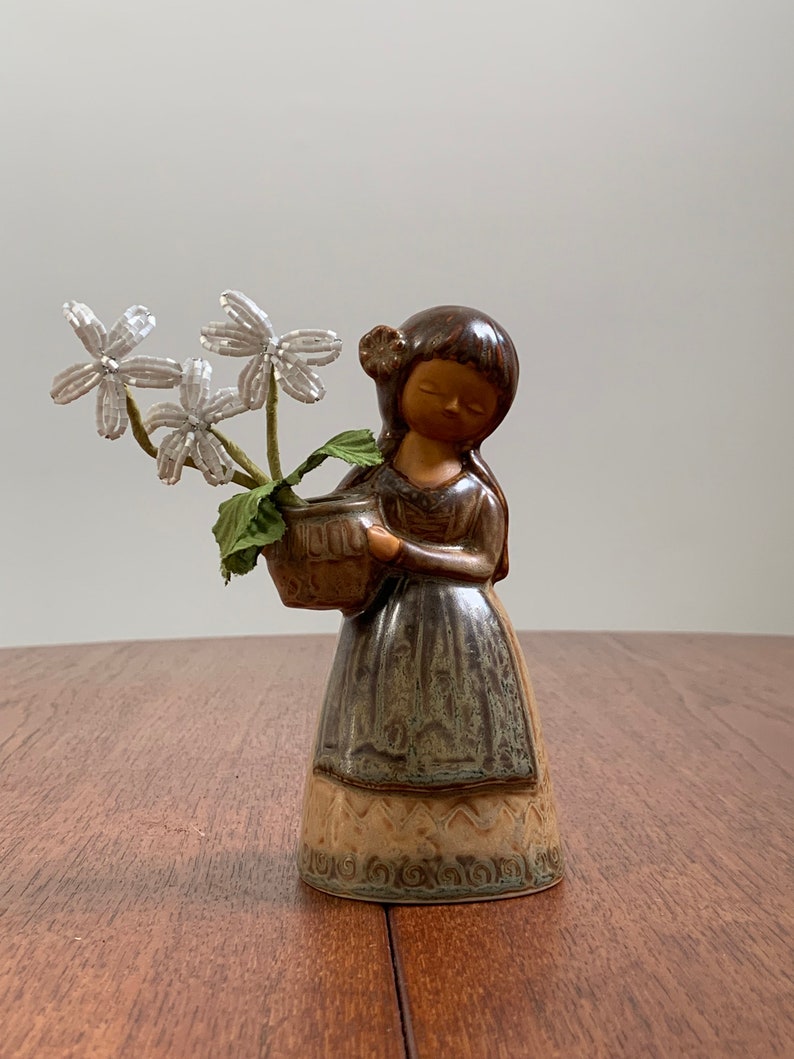 May include: A ceramic figurine of a girl holding a basket of white flowers. The girl is wearing a brown dress with a green and brown pattern. The figurine is made in a rustic style.