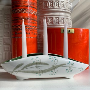 May include: White ceramic candle holder with a green rim and a floral design. Three white candles are in the holder.