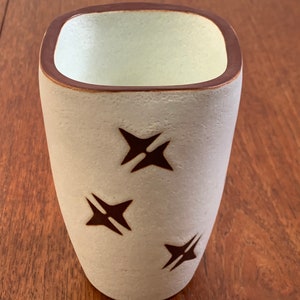 May include: A white ceramic cup with a brown rim and brown star designs. The cup is on a wooden surface.