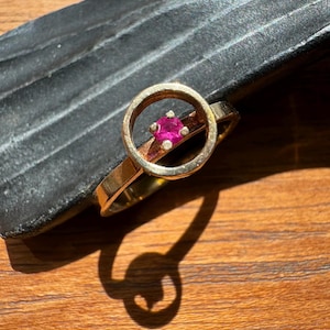 Peut inclure: Une bague dorée présentant un design à cadre circulaire avec une seule petite pierre précieuse ronde d'un rose vif sertie au centre. L'anneau en métal a une finition lisse et l'élément circulaire est positionné horizontalement sur l'anneau.