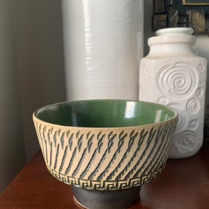 May include: A green and white ceramic bowl with a geometric design. The bowl has a wide rim and a narrow base.