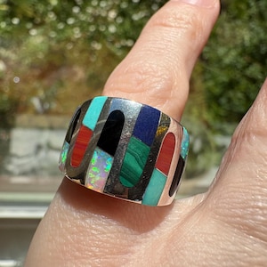 May include: A wide, silver ring with a mosaic design featuring various gemstones. The ring showcases rectangular inlays of turquoise, red, black, green, and blue stones, along with iridescent opal accents. The ring is approximately 1 inch wide.