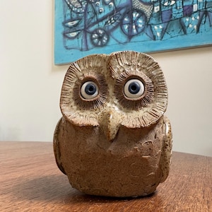 May include: A ceramic owl figurine with large, wide blue eyes and a textured, brown body. The owl has a detailed, feathered appearance and is sitting on a wooden surface. A blue and white abstract painting hangs in the background.