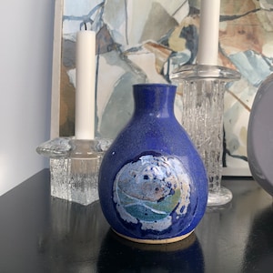 May include: A dark blue ceramic vase with a textured, abstract design in shades of blue, green, and beige. The vase has a rounded body and a narrow neck. The vase is on a black surface.