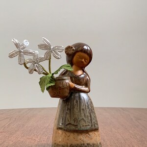 May include: A ceramic figurine of a girl holding a brown pot with white flowers. The girl is wearing a brown and green dress with a brown hair bun. The figurine is on a wooden surface.