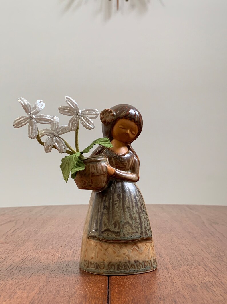 May include: A ceramic figurine of a girl holding a brown pot with white flowers. The girl is wearing a brown and green dress with a brown hair bun. The figurine is on a wooden surface.