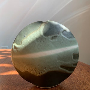 May include: A round ceramic vase with a textured, green and black glaze. The vase has a unique, organic shape and a white stripe around the middle.