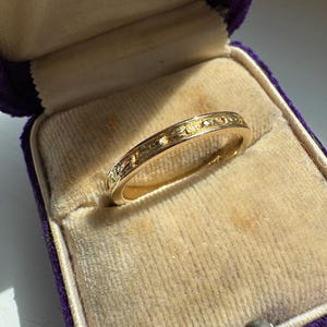 May include: A gold ring with a detailed pattern, presented in a vintage purple velvet ring box. The ring's surface features a repeating design. The box shows signs of age, with a worn, aged appearance.