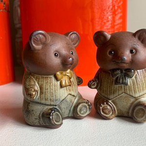 Może przedstawiać: Dwie brązowe ceramiczne figurki niedźwiedzi w brązowo-białych paskowanych strojach z żółtymi i czarnymi muszkami. Niedźwiedzie siedzą na białym tle.