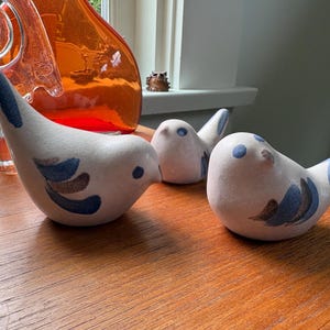 May include: Three ceramic bird figurines with a matte off-white finish. Each bird features hand-painted blue and brown wing details, a small blue eye, and a rounded body shape. The birds are arranged in a group, showcasing their smooth, curved forms.
