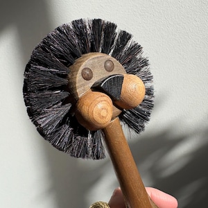 May include: A wooden toilet brush shaped like a lion's head. The brush has a dark brown handle and a circular head with black bristles. The lion's face is made of light brown wood with round eyes and a black nose.