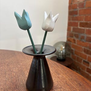 May include: A dark brown vase with two wooden tulip flowers. One is light blue, the other white. The vase has a wide, shallow top and a conical body. The flowers have green stems.