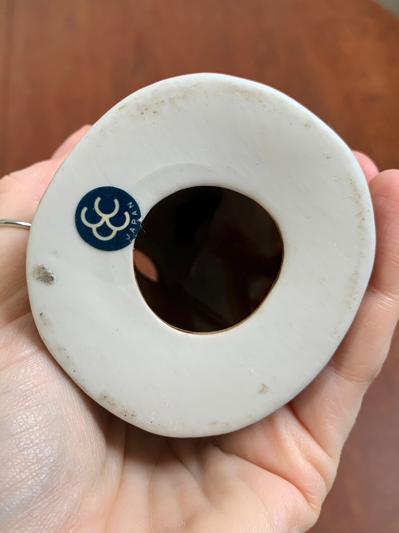 May include: White ceramic figurine with a blue and white sticker on the bottom that reads "JAPAN" and has a triple C logo. The figurine has a hole in the bottom.