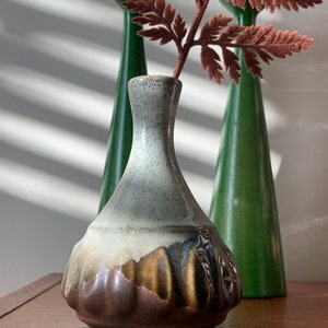 Op de afbeelding: Een kleine, grijze en bruine keramische vaas met een getextureerd, bolvormig lichaam. De vaas staat op een houten oppervlak met een bruine varen erachter.