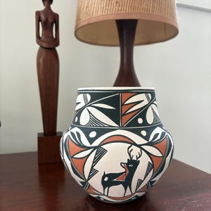 May include: A white ceramic pot with a black and orange geometric design featuring a deer. The pot is on a wooden table.