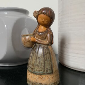 May include: A ceramic figurine of a girl holding a bowl. The figurine is brown and green with a detailed design.
