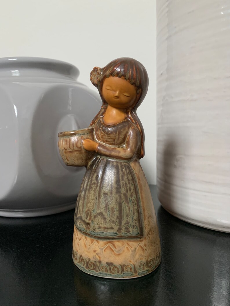 May include: A ceramic figurine of a girl holding a bowl. The figurine is brown and green with a detailed design.