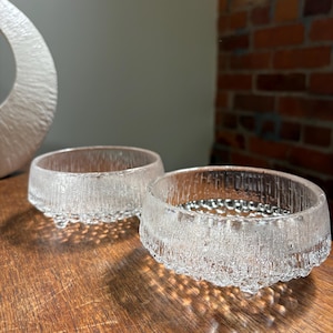 May include: Two clear glass bowls with a textured, frosted exterior finish. Each bowl features a vertical ribbed pattern along the sides and a series of small, rounded glass feet at the base, creating a distinct, tactile appearance for these vessels.