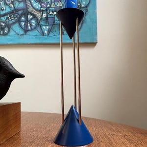 May include: A contemporary candle holder with a blue conical base and a black conical top. The holder has thin, gold-coloured metal rods and holds a blue twisted candle. The background includes a blue abstract painting.