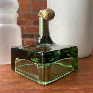 以下が含まれることがあります： 正方形の底と金色の金属製の蓋が付いた緑色のガラス瓶。瓶は木製の表面の上に置かれています。