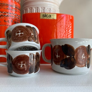 Peut inclure: Un ensemble de trois tasses en céramique brun et blanc avec un motif floral et un pot à crème assorti en céramique brun et blanc. Les tasses sont empilées les unes sur les autres. Le pot à crème est posé sur une surface blanche.