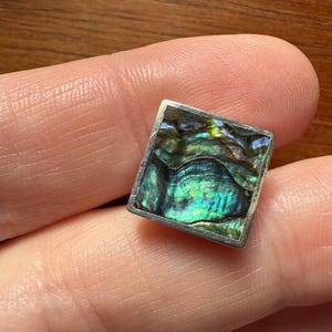May include: A square silver earring with an iridescent abalone shell inlay. The shell displays a mix of green, blue, and purple hues. The earring is held between two fingers, showcasing its size and design.