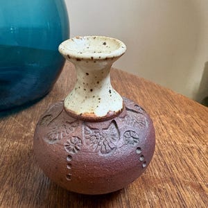 Puede incluir: Un pequeño jarrón de cerámica artesanal, con cuerpo redondo marrón rojizo y cuello crema moteado. El cuerpo presenta patrones florales y de hojas en relieve. El jarrón se exhibe sobre una superficie de madera, con un objeto de vidrio verde azulado al fondo.