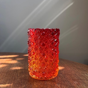 Könnte beinhalten: Ein zylindrisches Glas mit einem leuchtenden Farbverlauf von Rot zu Orange. Die Außenfläche ist mit einem erhabenen, sich wiederholenden geometrischen Sternmuster bedeckt, das kleine kreisförmige Akzente zwischen den Sternformen aufweist.