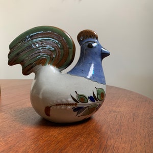 May include: A ceramic rooster figurine with blue, green and brown painted details. The rooster has a white body with a blue head and a green and brown tail.