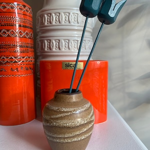 以下が含まれることがあります： 白と茶色のストライプ模様の小さな陶器の花瓶に、黒い木製のチューリップが2本挿してあります。花瓶は、オレンジと白の背の高い陶器の花瓶2つの前に、白い表面の上に置かれています。