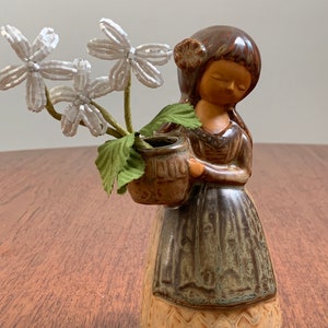 May include: A ceramic figurine of a girl holding a small vase with white flowers. The girl is wearing a brown dress with a green and brown pattern. The figurine is glazed in brown, green, and beige.