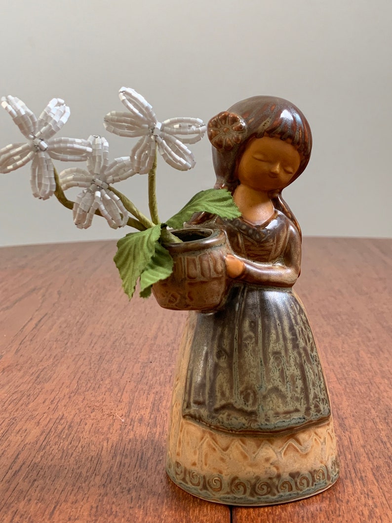 May include: A ceramic figurine of a girl holding a small vase with white flowers. The girl is wearing a brown dress with a green and brown pattern. The figurine is glazed in brown, green, and beige.
