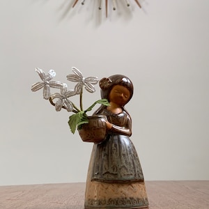May include: A ceramic figurine of a girl holding a brown pot with white flowers. The girl is wearing a brown dress with a green and brown pattern. The figurine is on a wooden table.